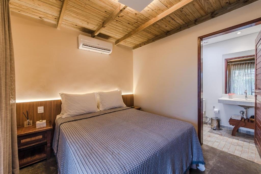 Quarto da Vila Salga Noronha, uma das pousadas em Fernando de Noronha, com cabeceira de madeira, mesinha com difusor, cama de casal com colcha cinza, ar-condicionado sobre a cama, e porta que dá para o banheiro na parede direita