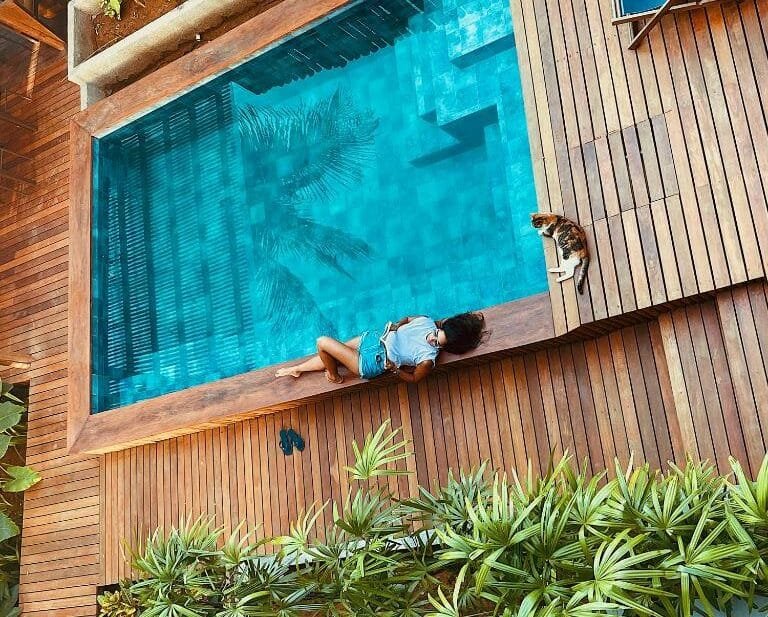 Piscina da Vila Salga Noronha, com pessoa deitada na beira, lendo livro, acompanhada de um gatinho malhado, que está deitado na beira adjacente