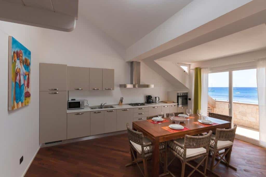 Parte do apartamento Villa ao Mar em Santa Maria que mostra uma mesa de madeira com cadeiras e ao fundo a cozinha com armários, aparelhos de cozinha e utensílios. Já do lado direito tem uma porta de vidro que dá acesso a varanda com vista para o mar. Imagem ilustrando post Hotéis em Cabo Verde.