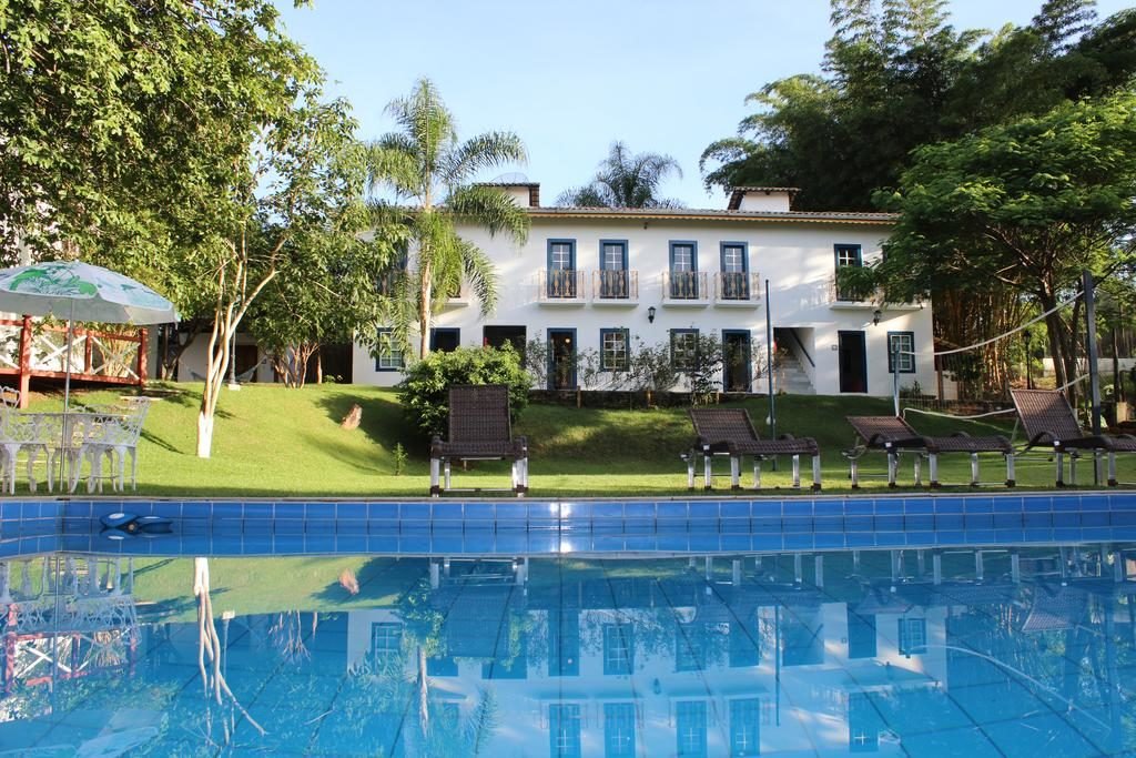 Área da piscina na Pousada Villa Bia em Pirenópolis, com cadeiras de praia, faixada da pousada e ao entorno vegetação. 