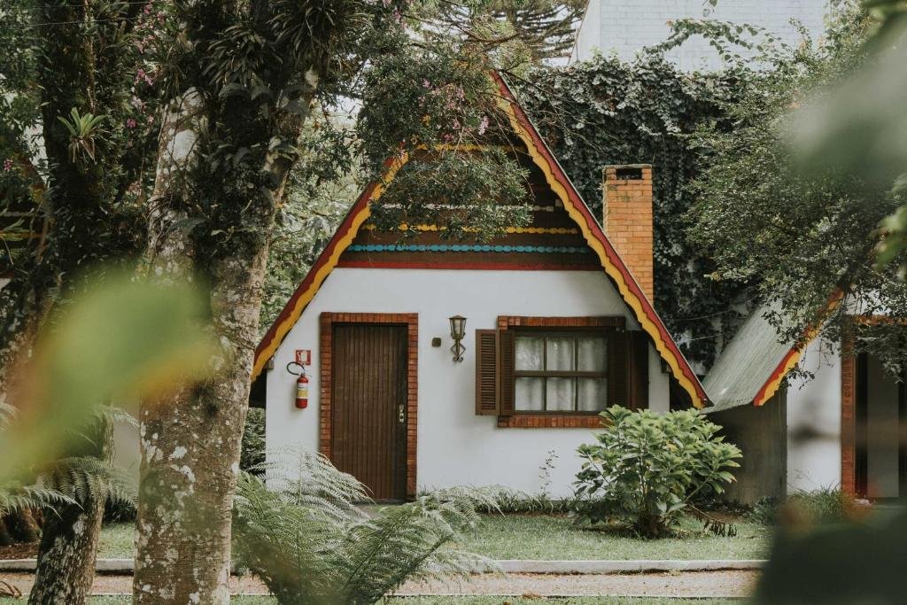 Exterior da Villa Chalés Gramado. Mostra a frente do chalé, que possui predes brancas, uma porta e janela de madeira e um telhado triangular. Por volta há uma vasta vegetação.