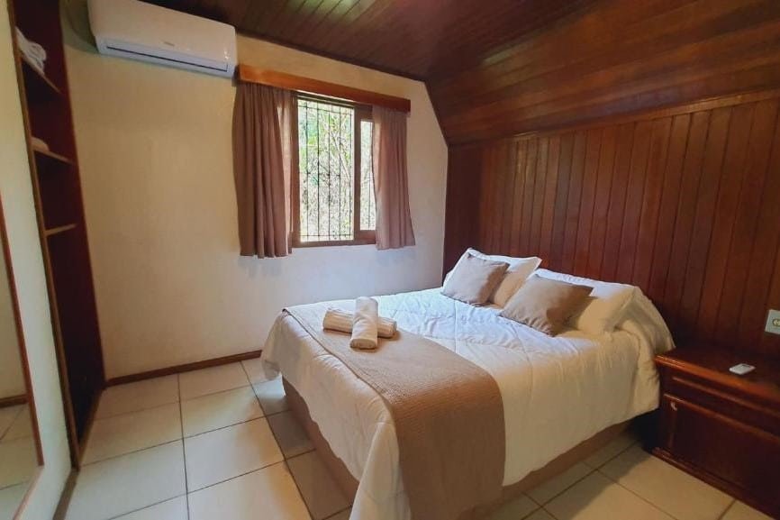 Foto do interior do Villa Chalés Gramado representando o post de chalés em Gramado. A imagem mostra um quarto com paredes e teto de madeira. No centro, há uma cama de casal com roupa de cama branca, dois travesseiros brancos e uma toalha dobrada em cima. À esquerda da cama, há uma pequena mesa de cabeceira de madeira sem itens visíveis. Acima da mesa, há um ar-condicionado montado na parede. À direita da cama, há uma janela com cortinas marrons parcialmente abertas, permitindo a entrada de luz natural.