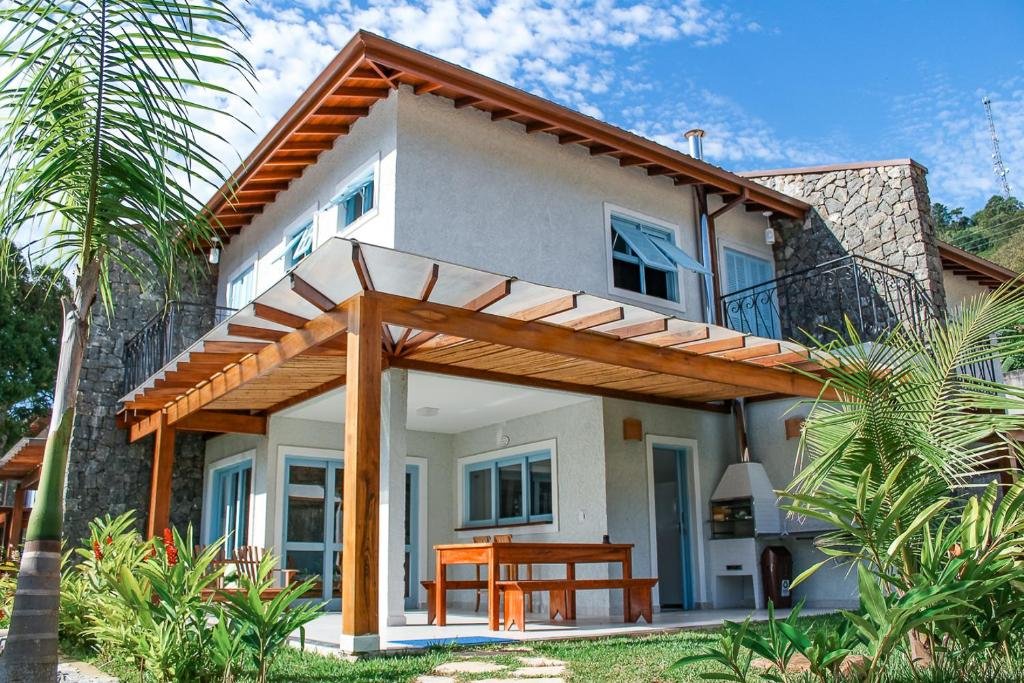 Casa do Villa Divina Pietra - Ubatuba com dois andares, pintada de cinza e com janelas azuis, na parte debaixo tem uma churrasqueira e uma mesa com bancos de madeira