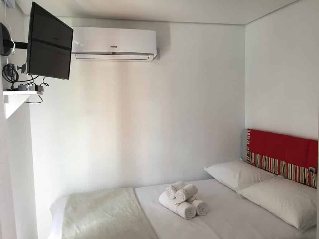 Quarto da Villa dos Ventos Hospedagem Container, uma das pousadas na Serra do Rio do Rastro. Uma cama de casal com toalhas dobradas em cima encara uma TV na parede oposta. Na parede lateral há um aparelho de ar-condicionado.