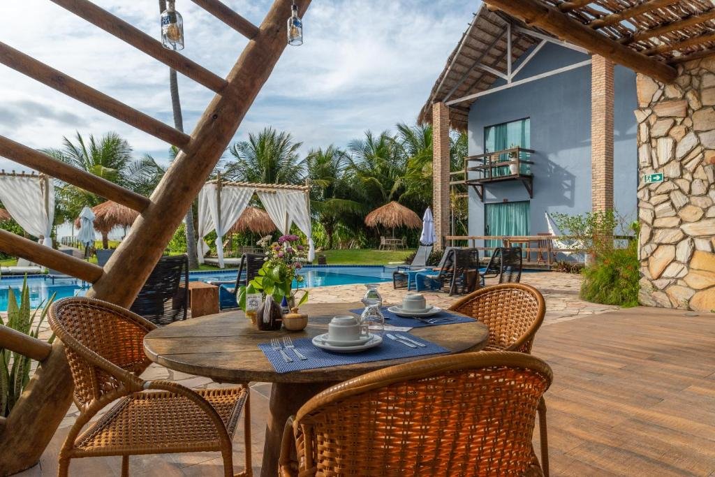 Área externa da Villa Pantai Milagres Exclusive Hotel, com uma mesa redonda em primeiro plano, mostrando lugares arrumados para duas pessoas, mas três cadeiras, e uma piscina ao fundo, com espreguiçadeiras e gramado mais atrás