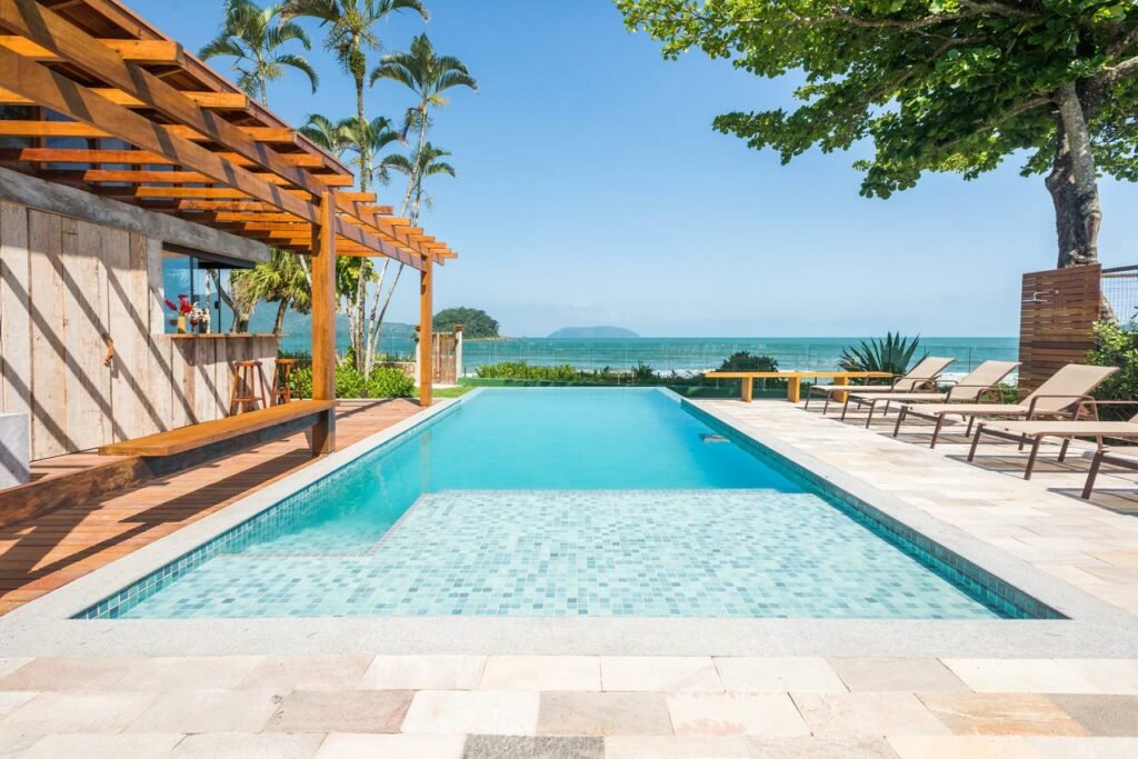 Piscina da Villa Sapê Pousada. Ela é retangular e está no meio da imagem. Ao seu lado esquerdo está o bar e ao lado direitos espreguiçadeiras. Ao fundo é possível ver o mar.