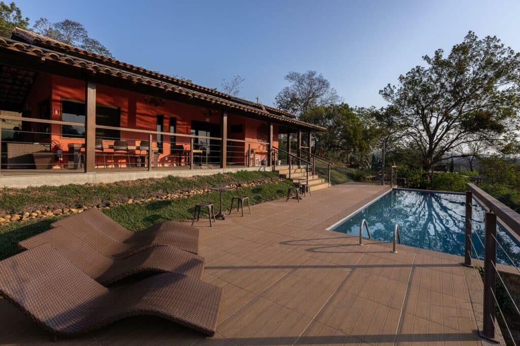Piscina da Villa Severina com bastante natureza no lado direito e algumas espreguiçadeiras no lado esquerdo. Há também a varanda da propriedade, onde tem mesa com cadeiras e a parede é na cor laranja, para ilustrar as pousadas em Macacos