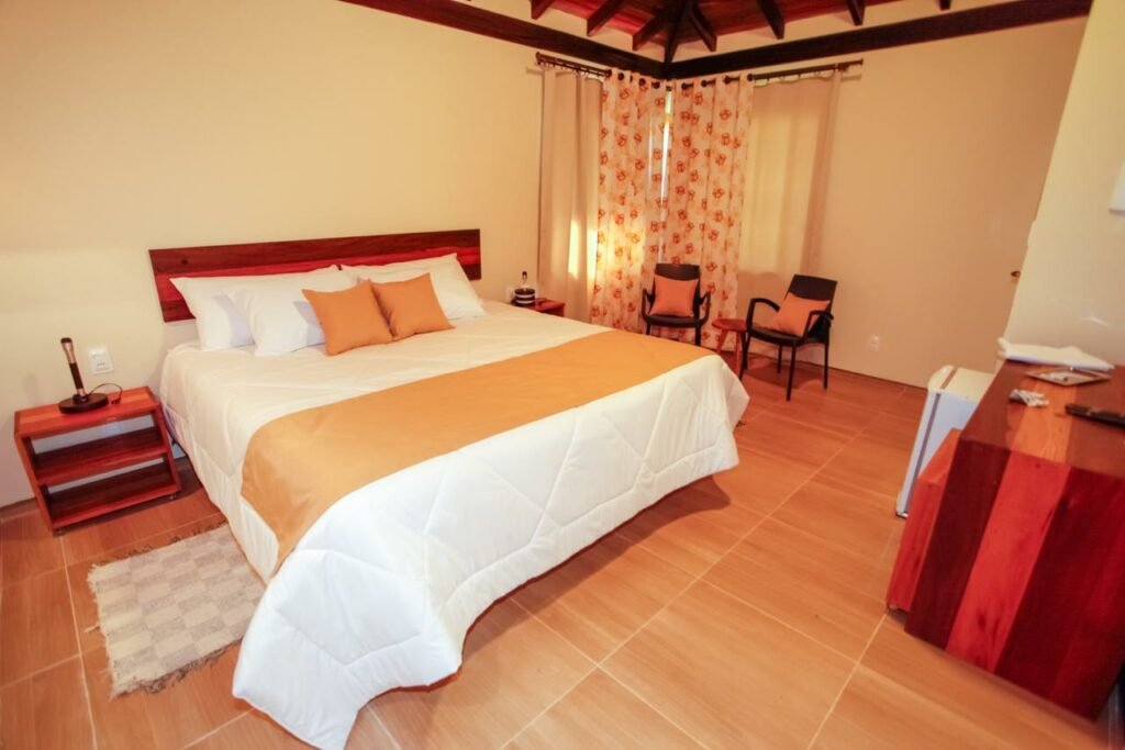 Quarto da Villa Severina, com cama de casal, duas cadeiras com uma almofada em cima de cada, duas escrivaninhas, sendo uma de cada lado da cama, e um armário pequeno com frigobar. A decoração do quarto usa os tons laranja e bege