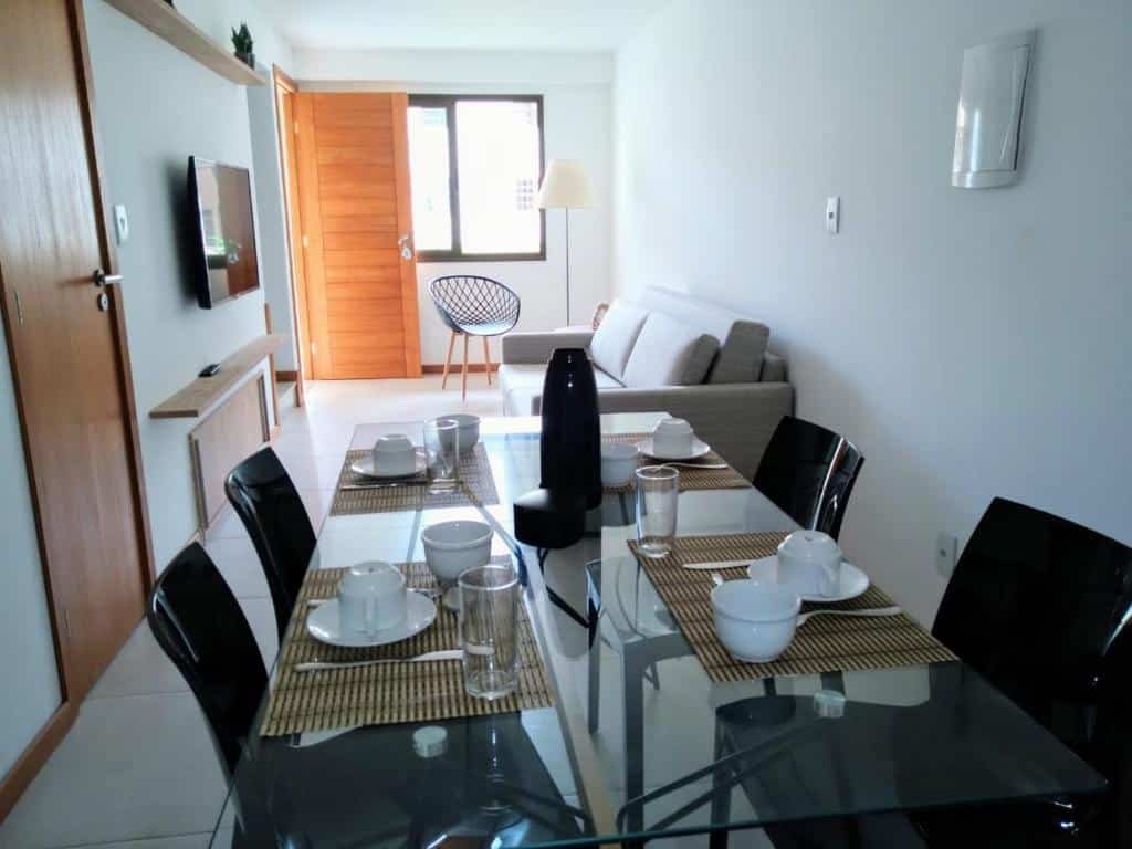 Sala de estar e de jantar na Village Angelim. Em primeiro plano vemos a mesa de jantar com quatro lugares, posta com xícaras e copos. No fundo está a sala. O sofá fica à esquerda e a TV na sua frente. Mais ao fundo  há uma cadeira, um abajur, uma janela e uma porta.