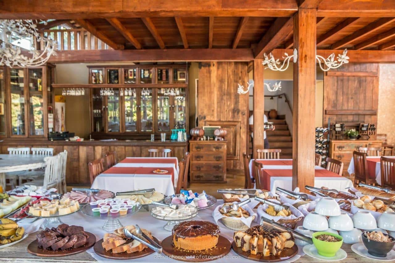 Um restaurante rústico com uma mesa de bufê exibindo diversas sobremesas e queijos, ao lado de mesas e cadeiras de madeira.