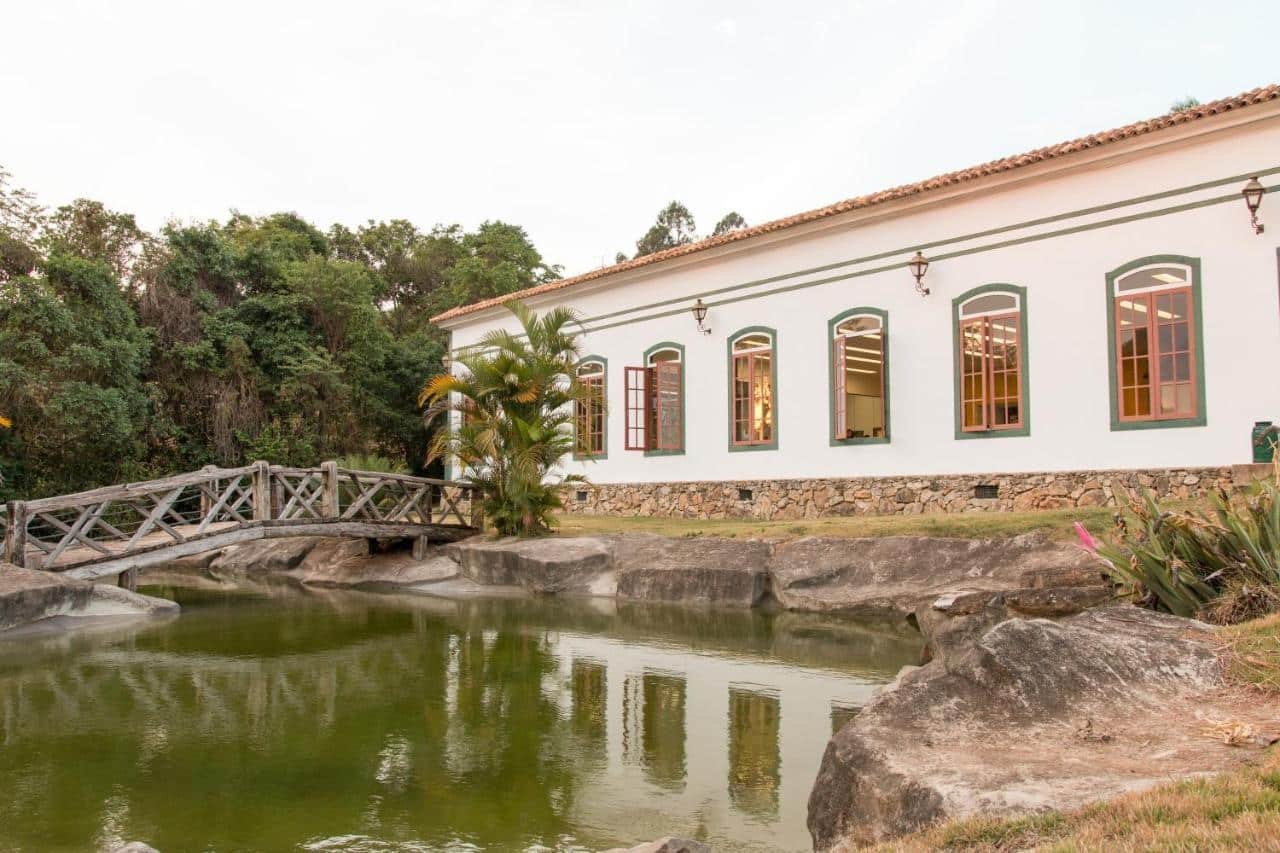 Edifício branco colonial do Ville Real Hotel, um dos hotéis fazenda em Minas Gerais, com janelas de madeira perto de um lago com ponte, cercado por pedras e vegetação.