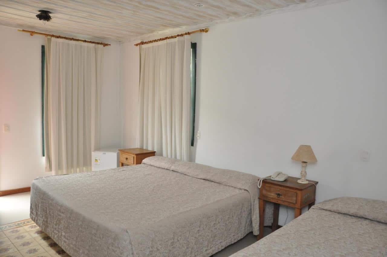 Um quarto com duas camas, duas mesas de cabeceira com lâmpadas, um telefone, cortinas brancas e colchas bege no Ville Real Hotel. Há uma pequena geladeira no canto.