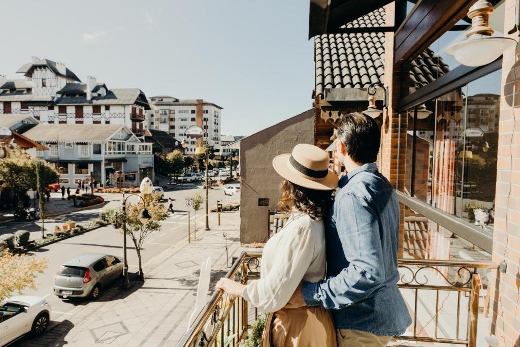 um casal abraçado, no lado direito da imagem, olhando para rua de Gramado de dentro de uma sacada aberta. Há carros na rua abaixo, no canto esquerdo inferior