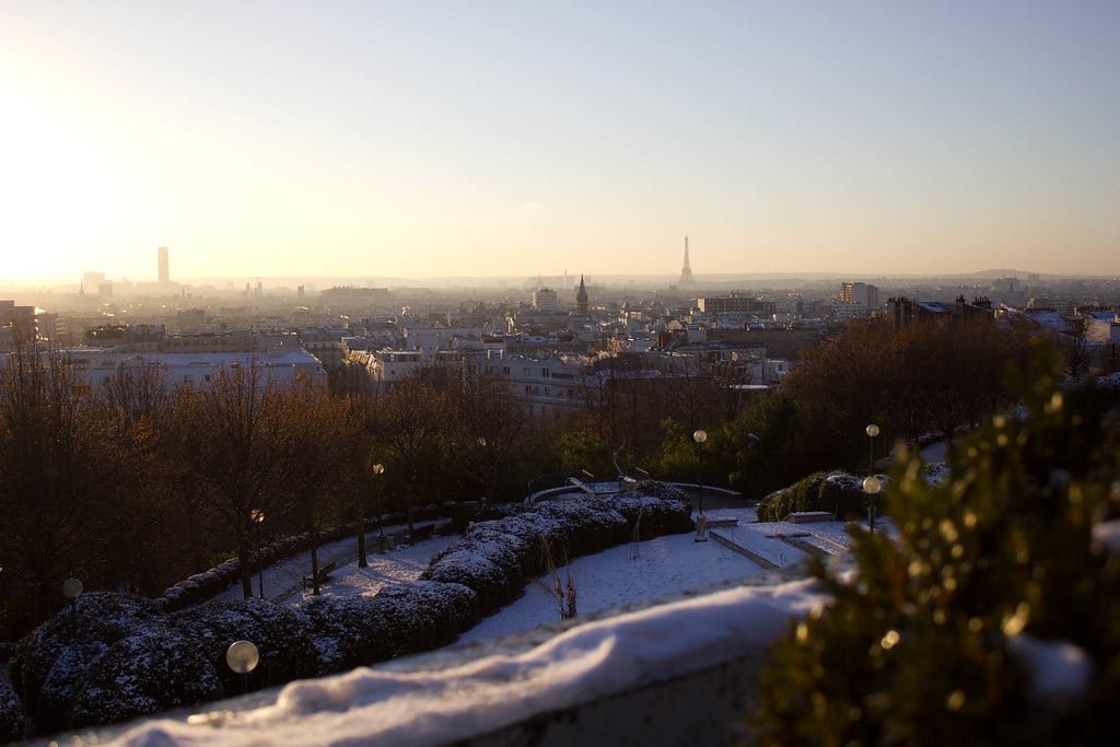 Uma paisagem urbana de Paris ao pôr do sol, com a Torre Eiffel ao longe e telhados cobertos de neve em primeiro plano. Essa é a vista do Parque de Belleville.