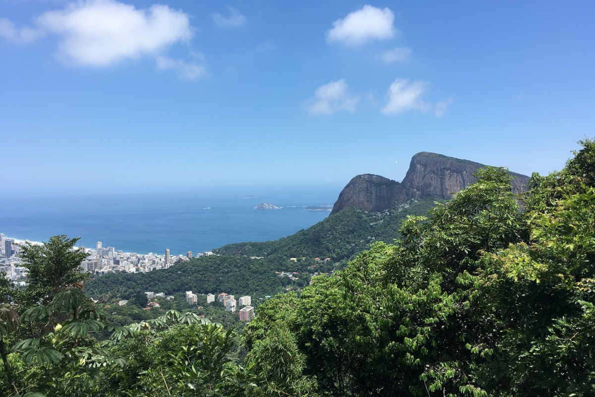 Vista de uma cidade costeira com altas montanhas verdes, um céu azul claro e vegetação exuberante em primeiro plano. Essa é a paisagem de uma das trilhas no Rio de Janeiro.