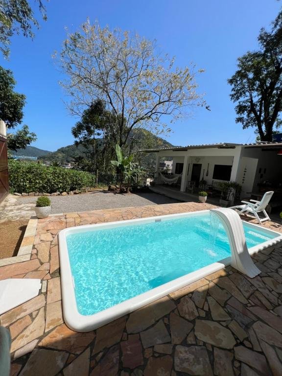 Área externa da Vista pro mar. Na frente uma piscina com cascata, do lado direito a casa, no fundo um jardim, árvores e vista para o mar.