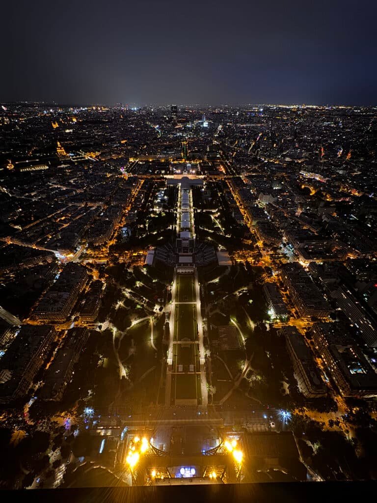 Vista aérea de Paris à noite, mostrando ruas e edifícios iluminados com uma estrutura iluminada de destaque no centro.