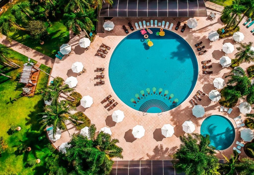 Imagem aérea do Vivaz Cataratas Hotel Resort que mostra uma piscina redonda grande e do lado uma piscina redonda pequena. Em volta das duas piscinas há vários guarda-sóis, espreguiçadeiras e árvores. Imagem ilustrando post Resorts em Foz do Iguaçu.