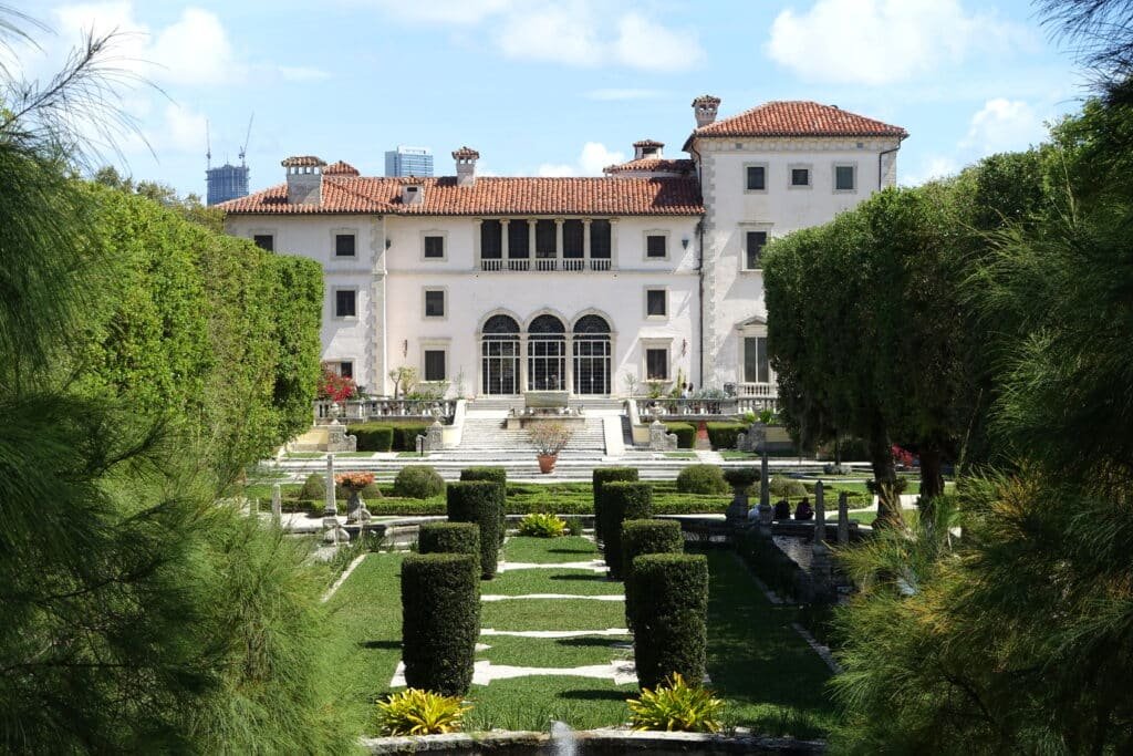 A imagem mostra uma pequena parte dos jardins no Vizcaya Museum and Gardens e a entrada da mansão. Os jardins são bem decorados e organizados. A casa é toda branca por fota, possui várias janelas, uma escada até as portas e vários andares. Essa é uma opção mais cultural de o que fazer em Miami.