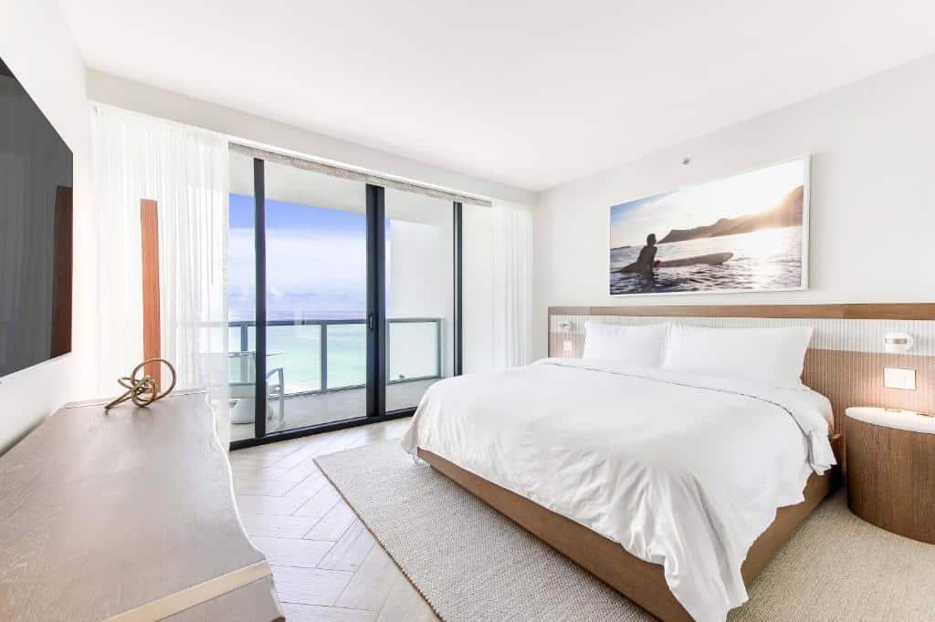 A imagem mostra um quarto decorado em tons claros no W South Beach. Há uma cama de casal grande com roupa de cama branca e uma cabeceira de madeira. Na parede acima da cama, há uma grande foto de uma pessoa com uma prancha de surfe no mar. O quarto tem uma grande porta de vidro que leva a uma varanda com vista para o oceano, deixando entrar muita luz natural. À esquerda da cama, há uma cômoda com uma decoração minimalista. Cortinas brancas leves emolduram a vista para o mar, criando uma atmosfera serena e relaxante.