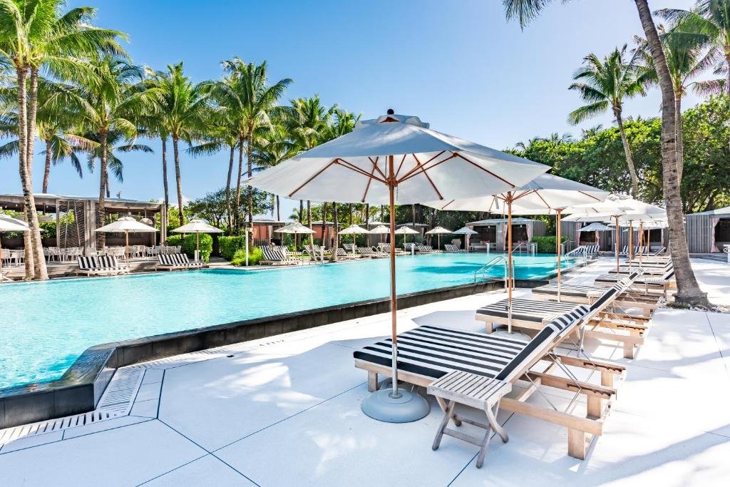 A imagem mostra uma área de piscina ao ar livre cercada por palmeiras altas no W South Beach, um dos hotéis em Miami Beach indicados. A piscina é grande e retangular, com água clara e azul. Ao redor da piscina, há muitas espreguiçadeiras listradas de branco e preto, cada uma sob um guarda-sol branco. O chão ao redor da piscina é de concreto branco, que reflete a luz do sol. Ao fundo, há mais espreguiçadeiras e áreas de descanso à sombra de árvores e plantas tropicais, criando um ambiente relaxante e tropical.