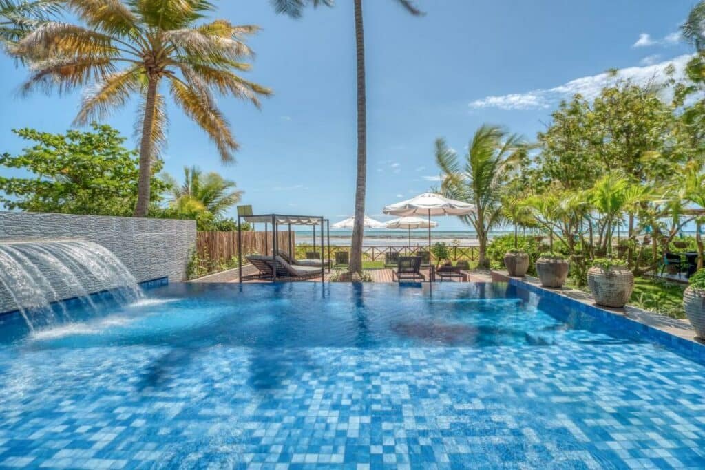 Piscina com cascata na Wassu Pousada, sendo o chão todo em mosaico de diferentes tons de azul, à frente há espreguiçadeiras, e ao fundo já está a praia e o mar.