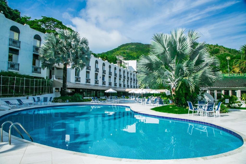 Piscina do Wembley Inn Hotel na Praia das Toninhas, um local amplo e com algumas árvores, cadeiras e mesas, espreguiçadeiras, guarda-sóis, do lado esquerdo é possível ver as sacadas do hotel e, mais ao fundo, as montanhas