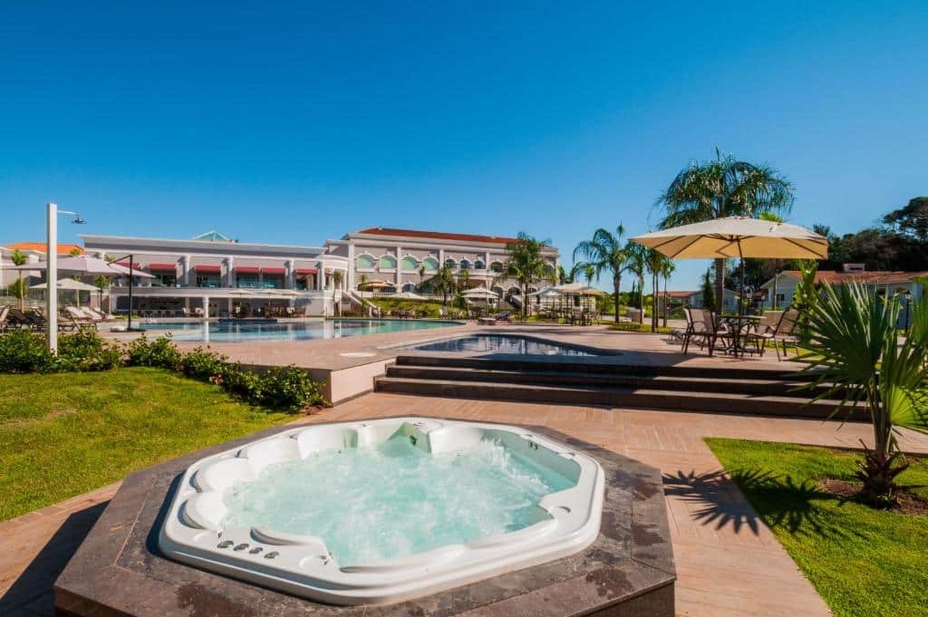 Parte do resort Wish em Foz do Iguaçu que mostra uma banheira de hidromassagem ao ar livre e ao fundo uma enorme piscina também ao ar livre. Em volta tem algumas árvores e gramado verde durante o dia. Imagem ilustrando post Resorts em Foz do Iguaçu.