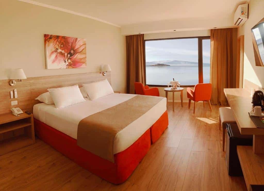 Quarto do Xelena Hotel & Suites. Uma cama de casal no lado esquerdo, de cada lado uma mesa de cabeceira e um abajur. De frente uma mesa, com um banco estofado, um frigobar e televisão. No fundo do quarto duas cadeiras e uma mesinha redonda com champagne viradas para a janela com vista para o lago. Foto para ilustrar post sobre hotéis em El Calafate.