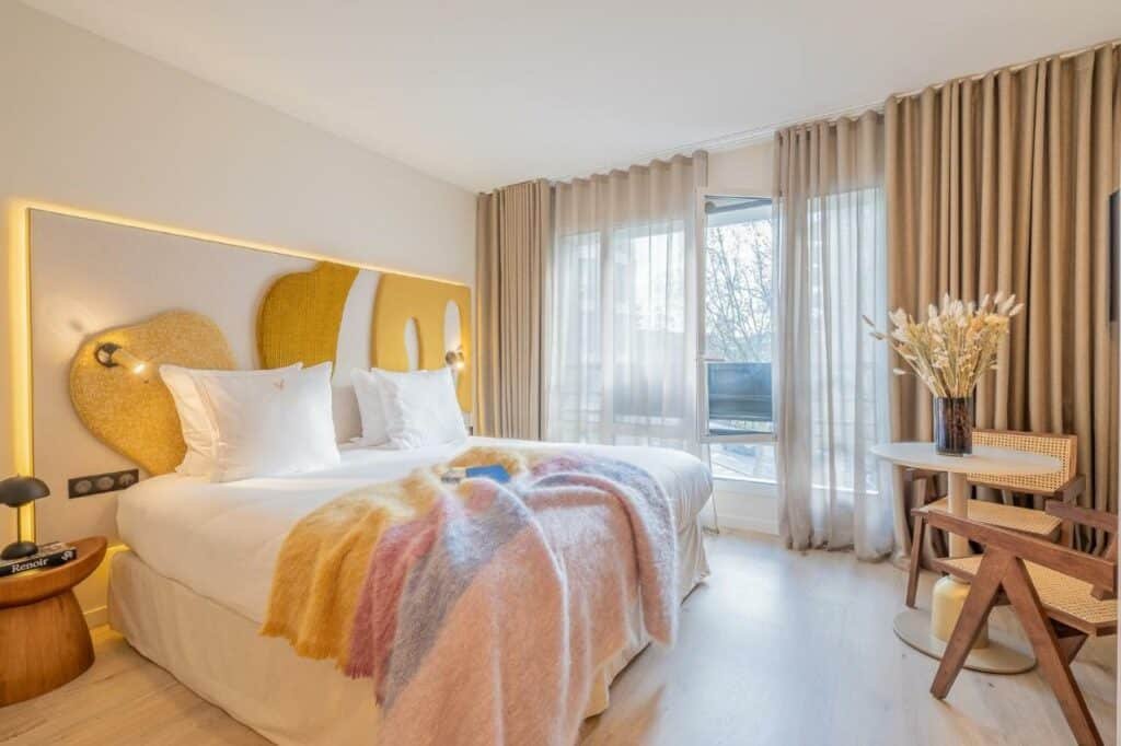 A imagem mostra um quarto elegante e bem iluminado no Yuna Porte-Maillot – ApartHotel. A cama grande tem lençóis brancos impecáveis e uma manta colorida, com um encosto de cabeceira em tons de amarelo e formas abstratas. Ao lado da cama, há uma mesinha de madeira com uma lâmpada moderna. Há uma janela grande com cortinas translúcidas e opacas, permitindo a entrada de luz natural. Perto da janela, há uma pequena mesa redonda com um vaso de flores secas e uma cadeira de madeira. O quarto é decorado em tons suaves, criando um ambiente acolhedor e tranquilo.
