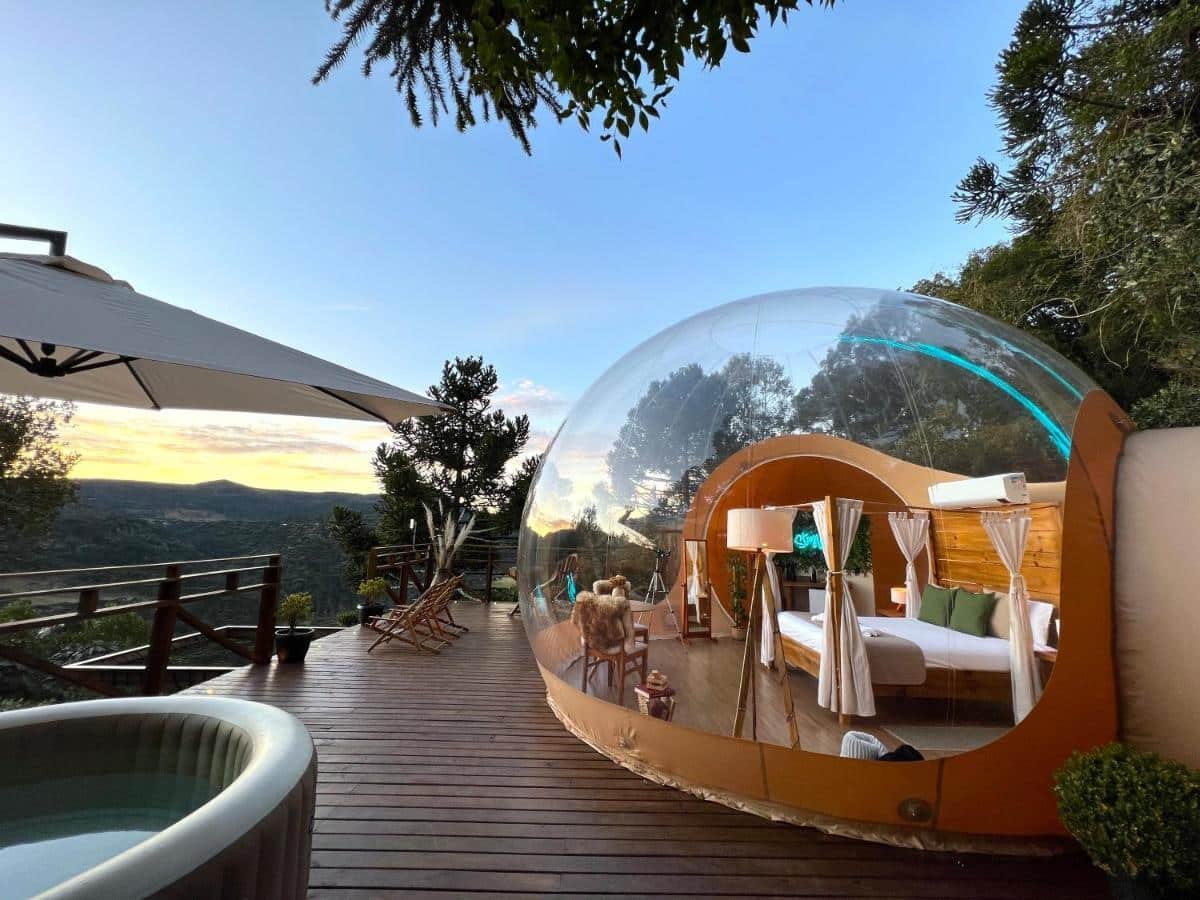 Vista geral do quarto e varanda do hotel Zion Bubble Glamping. O quarto está dentro de uma bolha transparente, que possibilita ver o céu e toda a paisagem do local. Uma cama de casal com dossel está no meio e algumas coisas como uma cadeira, espelho e um telescópio estão ao redor. Um ar-condicionado está acima da cama. Fora da bolha é possíver ver uma jacuzzi com guarda-sol, duas cadeiras de madeira e a vegetação como paisagem.